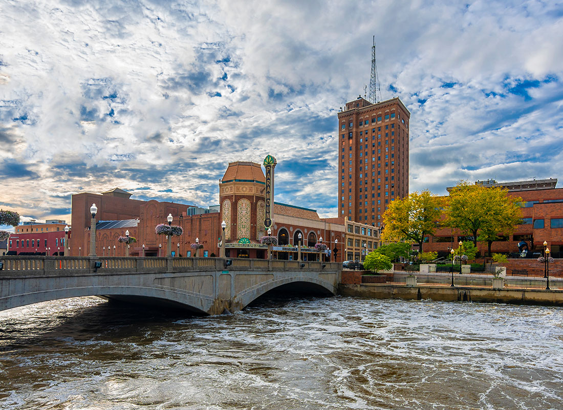 Aurora, IL - View of the Downtown Area in Aurora Illinois with Commercial Buildings Next to a Walking Bridge Over the River Against a Cloudy Blue Sky