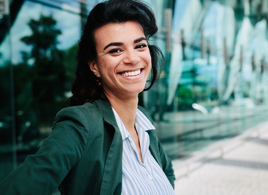 Service Center - Closeup Portrait of a Cheerful Young Business Woman Wearing a Button Down Shirt and Blazer Standing Outside a Modern Commercial Building in the City